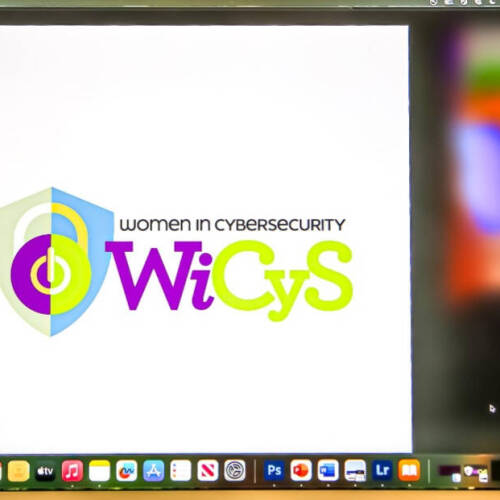 A computer screen displays the "Women in CyberSecurity (WiCyS)" logo, featuring a shield graphic and the text "WiCyS" in purple and lime green letters. The screen shows a Mac desktop with various app icons along the bottom